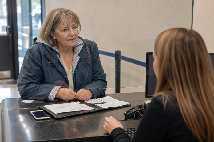 Older woman at a bank counter speaking with a cashier, a folder of printed receipts and a phone on the counter between them, daylight through the window