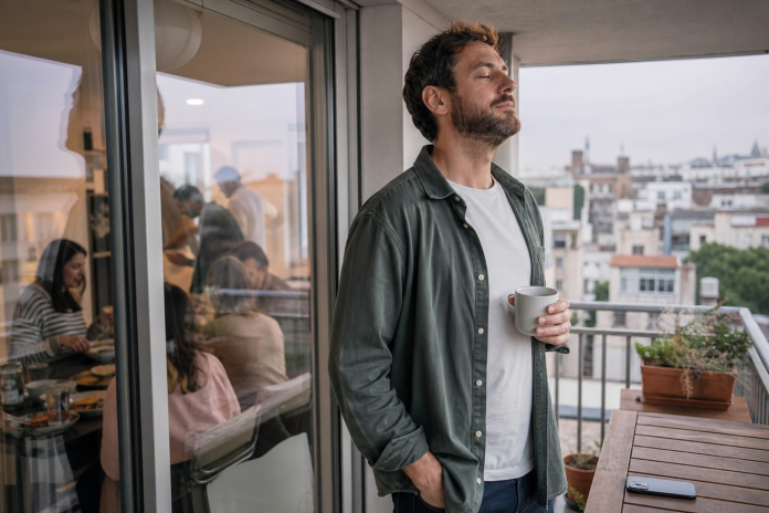 Man taking a breath on an apartment balcony while a family gathering continues inside
