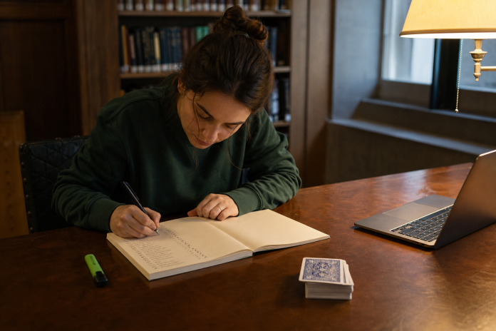 Woman at a library desk keeping a handwritten prediction journal next to a face-down deck of cards